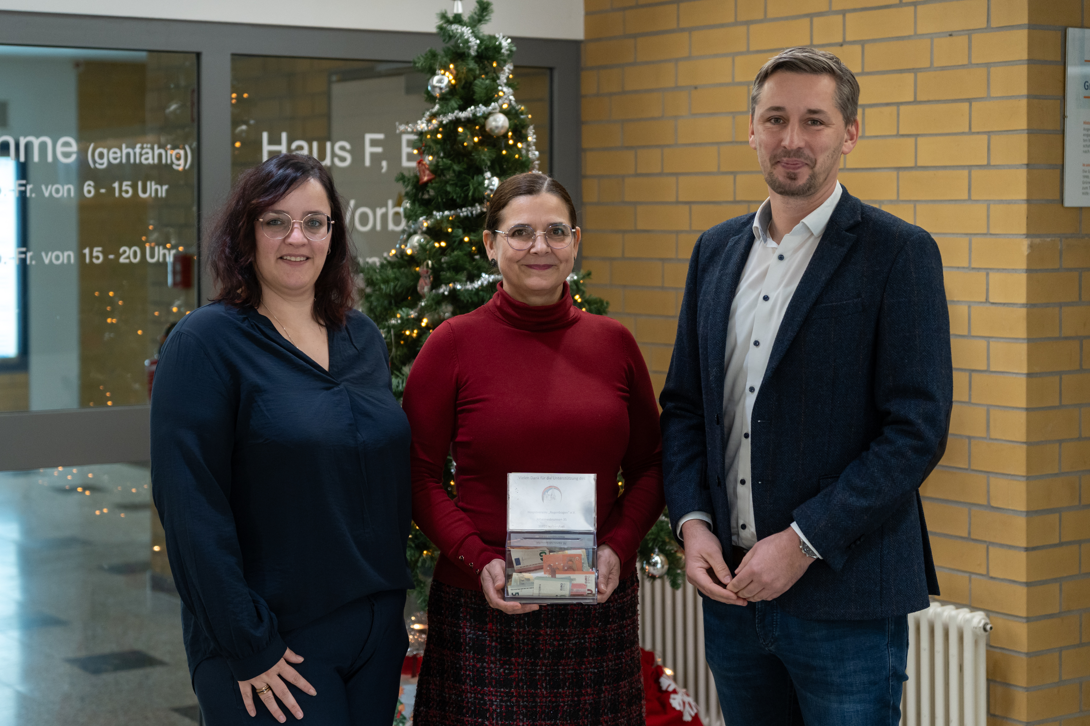 Zwei Frauen und ein Mann stehen vor einem Weihnachtsbaum und lächeln in die Kamera. Die Frau in der Mitte hält eine kleine Box mit Geld in der Hand.