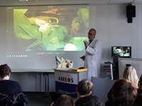 Arzt in Kittel steht vor einer Klasse und erklärt etwas anhand eines Ohr-Modell. Im Hintergrund sieht man eine Leinwand, auf der eine OP übertragen wird.