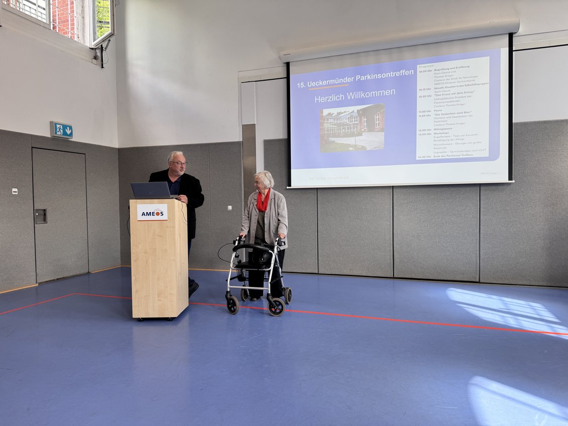 Thomas Krüger, Chefarzt der Klinik für Neurologie steht am Pult, rechts von ihm mit Rollator steht Karin Glause, die seit fast 30 Jahren die Parkinsonselbsthilfegruppen in MV anleitet und berät