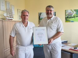 Zwei Ärzte in weißen Polo-Shirts stehen in einem Büro und halten lächelnd die Urkunde zur Rezertifizierung des Hypertonie-Zentrums der Deutschen Hochdruckliga e. V. DHL® in den Händen.